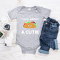 Grey baby romper/ onesie that has white writing that says Taco About A Cutie. To the middle is a smiling Taco.