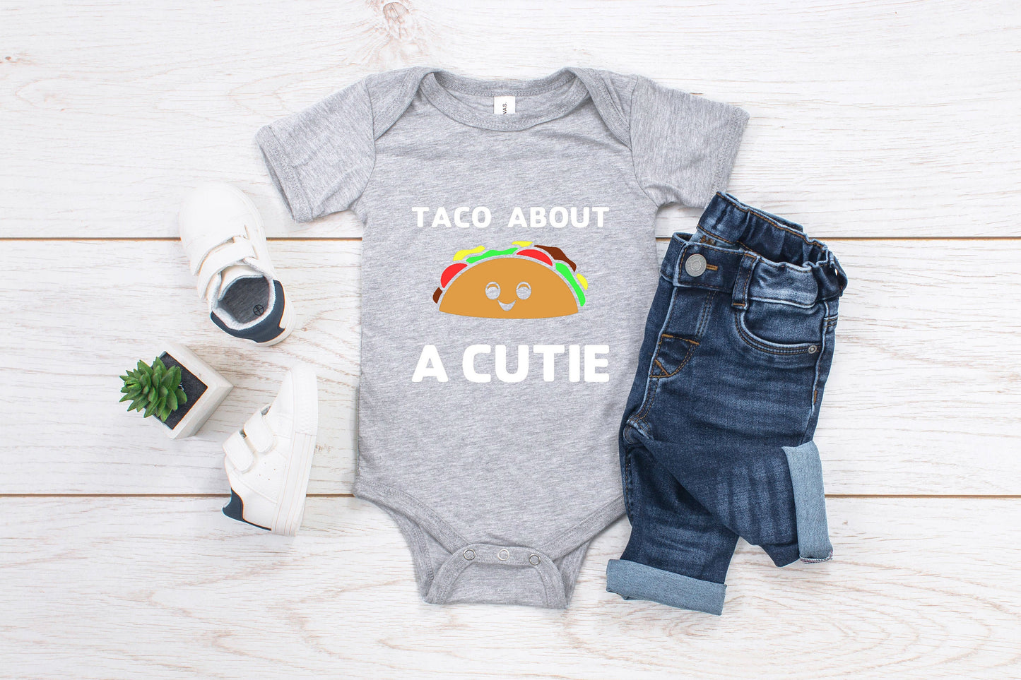 Grey baby romper/ onesie that has white writing that says Taco About A Cutie. To the middle is a smiling Taco.