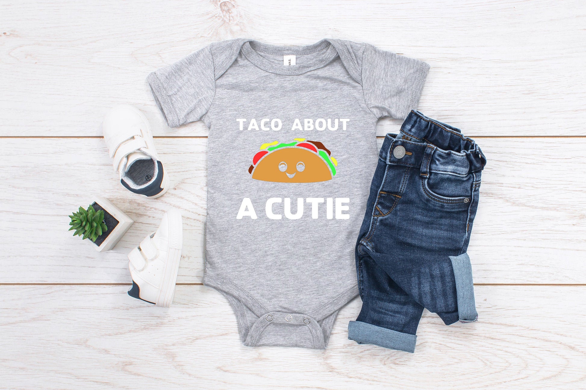 Grey baby romper/ onesie that has white writing that says Taco About A Cutie. To the middle is a smiling Taco.