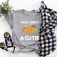 Grey t-shirt with white writing that says Taco About A Cutie. In between the writing is a smiling brown taco with brown, red, yellow and green coming out of it.