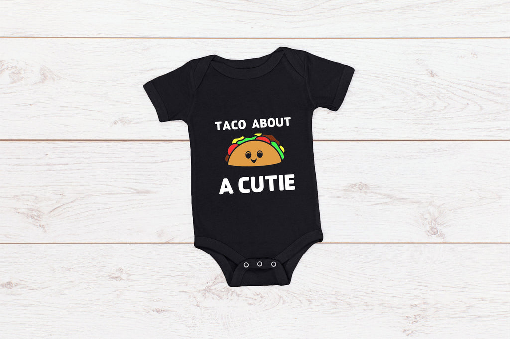 Black baby onesie with white writing that says Taco About A Cutie with a smiling Taco