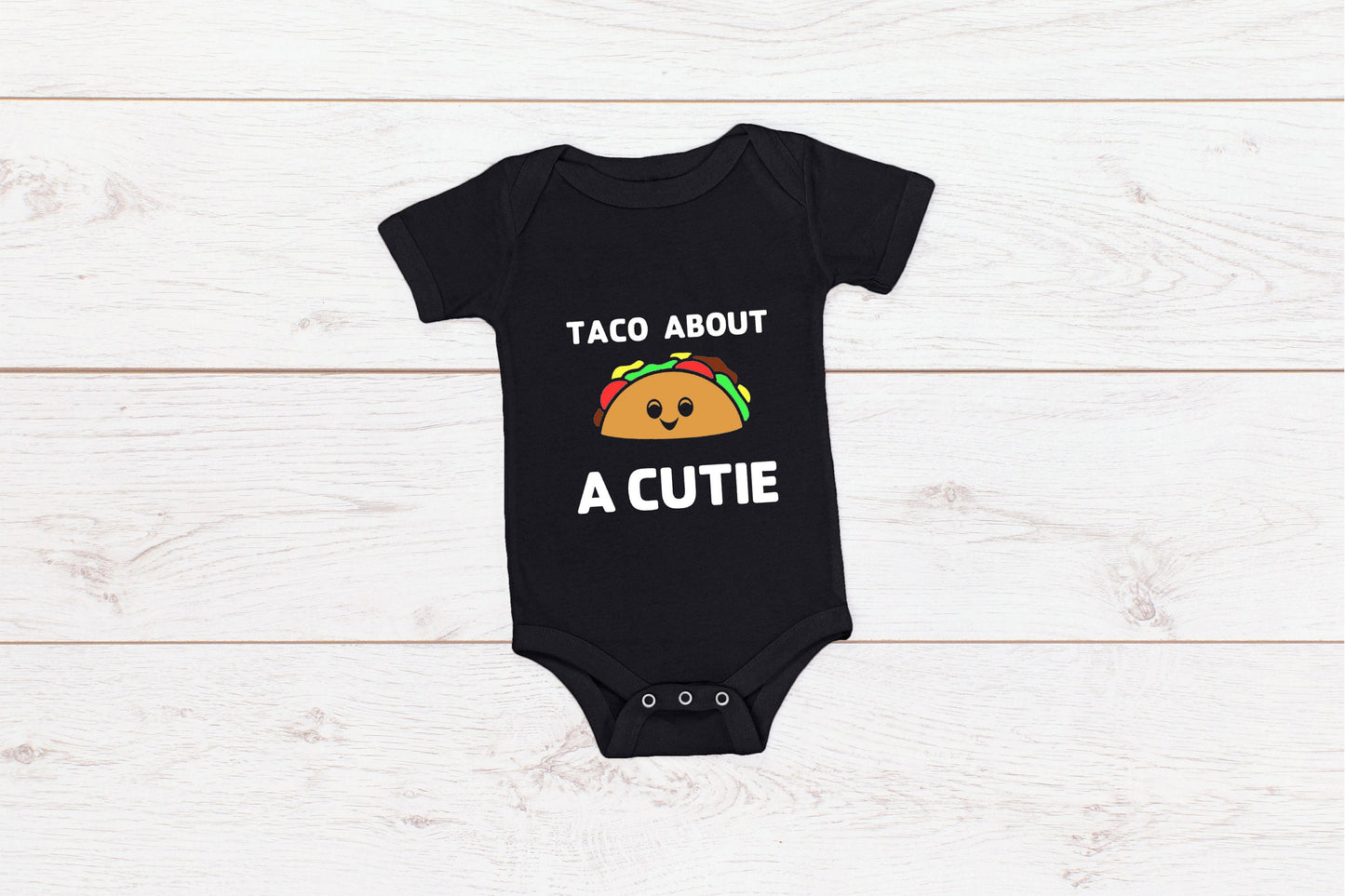 Black baby onesie with white writing that says Taco About A Cutie with a smiling Taco