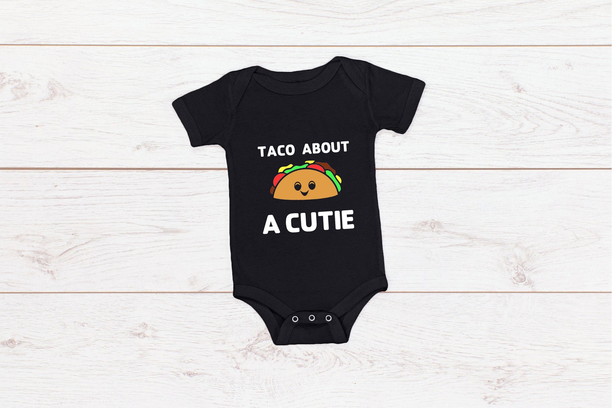 Black baby onesie with white writing that says Taco About A Cutie with a smiling Taco