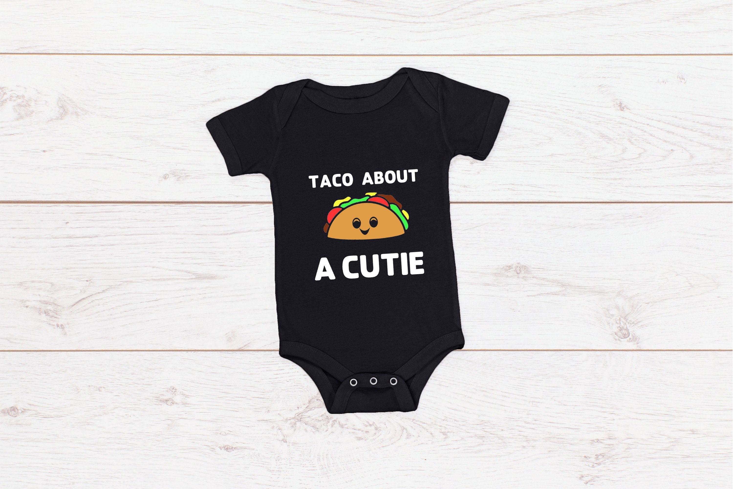 Black baby onesie with white writing that says Taco About A Cutie with a smiling Taco